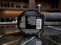 The back of a black circular cosmetic jar lid, showing white text with ingredients, a barcode, and a small red logo, resting on a dark marble surface with a blurred bathroom or vanity background.