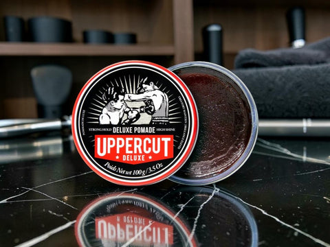 An open round tin of Uppercut Deluxe Pomade sits on a glossy black marble surface, with the lid leaning in front to show the label. The pomade inside is a dark reddish-brown.
