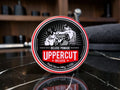 A round tin of Uppercut Deluxe Pomade sits on a glossy black marble surface with a blurred bar or salon background. The label features a black, white, and red design with an illustration of two men combing hair.