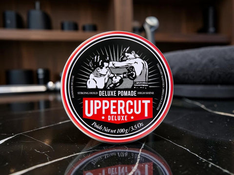 A round tin of Uppercut Deluxe Pomade sits on a glossy black marble surface with a blurred bar or salon background. The label features a black, white, and red design with an illustration of two men combing hair.