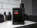 A bottle of Uppercut 3 in 1 Wash for hair, face, and body, standing on a bathroom counter with towels and other bottles in the background.