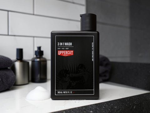 A bottle of Uppercut 3 in 1 Wash for hair, face, and body, standing on a bathroom counter with towels and other bottles in the background.