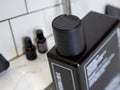 A close-up of a black rectangular bottle with a matching black cylindrical cap, photographed on a bathroom counter with white subway tiles and small dark bottles blurred in the background.