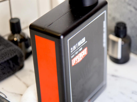 A bottle of UpperCut 3 in 1 Wash hair cleanser standing on a marble countertop with bathroom accessories blurred in the background.
