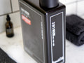 A black rectangular bottle of hair wash with white text and a red accent label, shown on a bathroom counter with a dark towel and small amber bottle in the background.