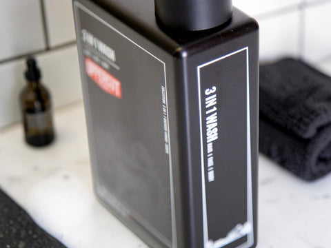 A black rectangular bottle of hair wash with white text and a red accent label, shown on a bathroom counter with a dark towel and small amber bottle in the background.