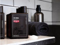 A bottle of Uppercut Men 3 in 1 Wash shampoo and conditioner set on a bathroom counter next to a rolled dark towel and soap dish, with a blurred background of white subway tiles.