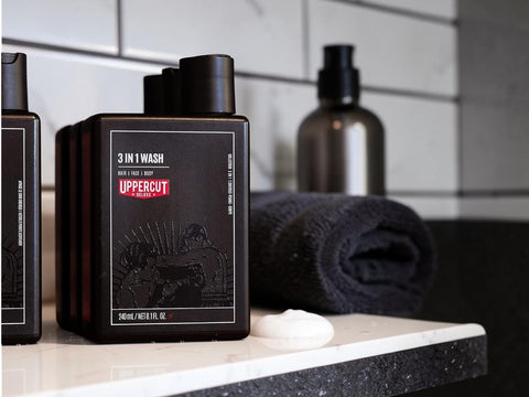 A bottle of Uppercut Men 3 in 1 Wash shampoo and conditioner set on a bathroom counter next to a rolled dark towel and soap dish, with a blurred background of white subway tiles.