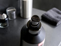A close-up of a matte black pump bottle with a black dispenser cap, set on a dark gray surface with blurred cosmetic jars and a folded dark towel in the background.