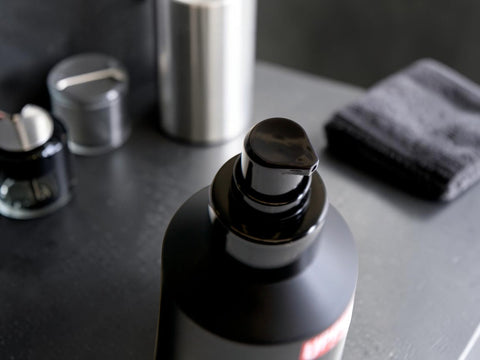 A close-up of a matte black pump bottle with a black dispenser cap, set on a dark gray surface with blurred cosmetic jars and a folded dark towel in the background.