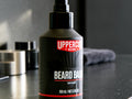 A bottle of Uppercut Deluxe Beard Balm with a black pump dispenser and a black label featuring a red and white logo, set on a dark countertop with blurred grooming items in the background.