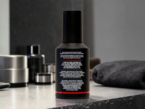 A black cylindrical bottle with a matching black cap stands on a dark countertop, showing the back label with white text and a thin red accent band. A folded dark towel and other containers are blurred in the background.