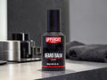 A 100 ml bottle of Uppercut Deluxe Beard Balm stands on a dark countertop in a modern bathroom setting, with a folded dark towel and other grooming items blurred in the background.