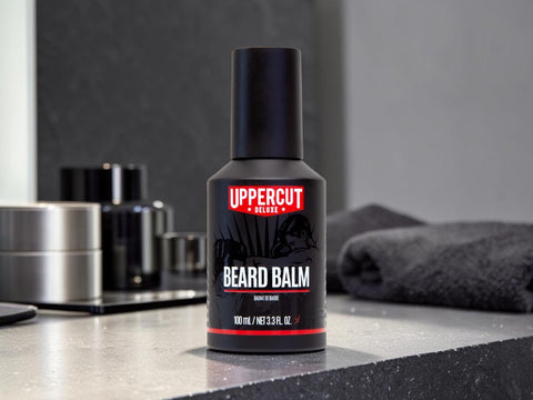 A 100 ml bottle of Uppercut Deluxe Beard Balm stands on a dark countertop in a modern bathroom setting, with a folded dark towel and other grooming items blurred in the background.