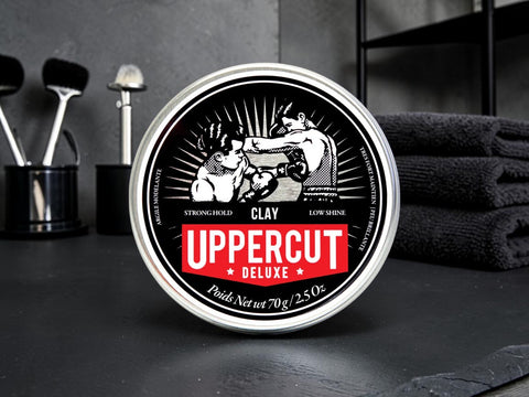 A round tin of Uppercut Deluxe Clay hair wax sits on a dark countertop with grooming tools and folded towels blurred in the background. The label is black and white with a bold red banner reading 'UPPERCUT DELUXE'.