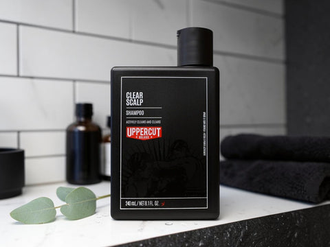 A black rectangular bottle of Uppercut Clear Scalp Shampoo stands on a bathroom counter with white subway tile behind it. A folded dark towel, a small green leaf sprig, and other dark bottles are visible in the background.