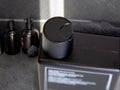 A matte black cylindrical bottle with a matching screw-top lid sits on top of a black rectangular box. The background shows a dark gray countertop with other dark bottles and a towel.