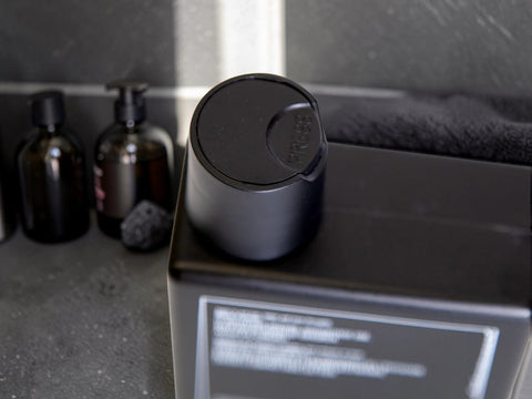 A matte black cylindrical bottle with a matching screw-top lid sits on top of a black rectangular box. The background shows a dark gray countertop with other dark bottles and a towel.