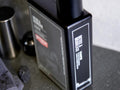 A close-up of a dark rectangular bottle labeled 'DETOX & SMOOTH' with a black cap, placed on a gray countertop with a towel and metal container nearby.
