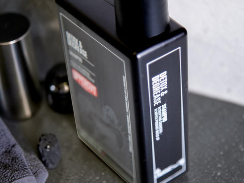 A close-up of a dark rectangular bottle labeled 'DETOX & SMOOTH' with a black cap, placed on a gray countertop with a towel and metal container nearby.