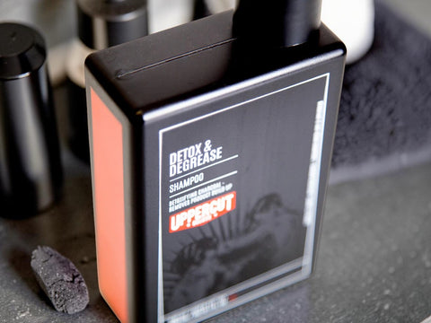 A bottle of UpperCut Detox & Degrease Shampoo standing on a dark countertop, with other dark bottles blurred in the background.