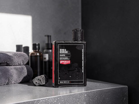 A bottle of Uppercut Detox & Regenerate Shampoo stands on a dark countertop beside folded gray towels and dark grooming bottles in a bathroom setting.