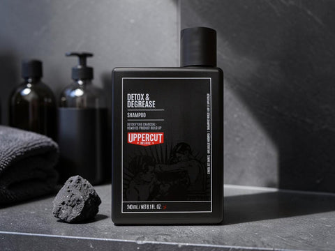 A matte black bottle of Uppercut London Detox & Degrease Shampoo stands on a dark stone surface with a folded gray towel and a small rock beside it. Two dark bottles and a soap dish are blurred in the background against a dark gray wall.