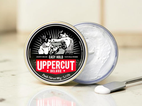 An open round tin of Uppercut Deluxe hair wax and a small spatula with white wax cream, set on a light marble-like surface with a blurred indoor background.