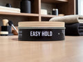 A black cylindrical hair product container labeled 'EASY HOLD' sits on a light wood surface with folded towels and grooming tools blurred in the background.