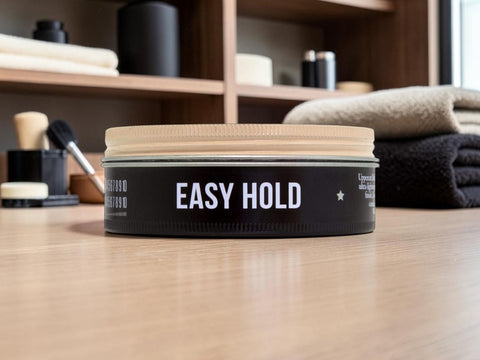 A black cylindrical hair product container labeled 'EASY HOLD' sits on a light wood surface with folded towels and grooming tools blurred in the background.