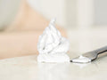 A dollop of thick white shaving cream or whipped body foam sits on a light marble surface next to a metal spatula, with a softly blurred warm-toned background.