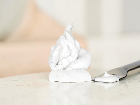 A dollop of thick white shaving cream or whipped body foam sits on a light marble surface next to a metal spatula, with a softly blurred warm-toned background.