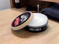 A round black tin of hair styling product labeled 'EASY HOLD' with its lid leaning against the container, set on a light wood surface. The lid label shows the brand 'PROFESSIONAL' with a red accent, and the product inside appears white or translucent.