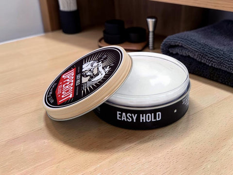 A round black tin of hair styling product labeled 'EASY HOLD' with its lid leaning against the container, set on a light wood surface. The lid label shows the brand 'PROFESSIONAL' with a red accent, and the product inside appears white or translucent.