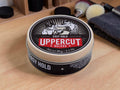 A round tin of Uppercut Deluxe Easy Hold hair styling product sits on a light wood surface, with grooming tools and a dark towel blurred in the background.