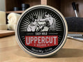 A round tin of Uppercut Deluxe Hair Hold styling product, featuring a vintage-style black and white illustration of a boxer on the lid, with a red banner reading 'UPPERCUT DELUXE'. The tin is placed on a wooden surface with a blurred background showing grooming items.