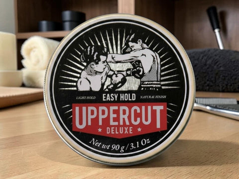 A round tin of Uppercut Deluxe Hair Hold styling product, featuring a vintage-style black and white illustration of a boxer on the lid, with a red banner reading 'UPPERCUT DELUXE'. The tin is placed on a wooden surface with a blurred background showing grooming items.