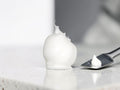 A dollop of thick white cosmetic cream or gel sits on a light stone surface beside a metal spatula holding a smaller amount, with a softly blurred gray background.