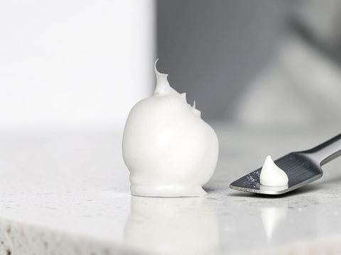 A dollop of thick white cosmetic cream or gel sits on a light stone surface beside a metal spatula holding a smaller amount, with a softly blurred gray background.
