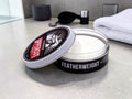 A round metal tin of Featherweight hair pomade, with the lid leaning against the open container to show the white pomade inside. The tin has a black label with white text and a red accent on the lid.