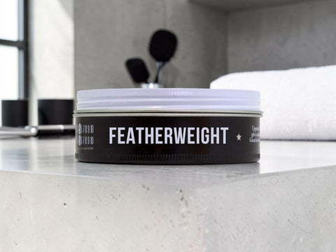 A tin of Featherweight hair pomade by T3D Beauty sits on a light gray stone countertop in a modern bathroom setting, with a white towel and black grooming tools blurred in the background.