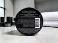 The back of a black circular hair product container, showing printed text, a barcode, and ingredients, photographed on a light gray countertop with blurred black containers in the background.