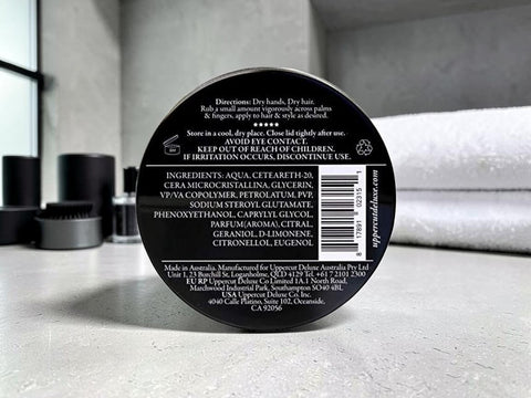 The back of a black circular hair product container, showing printed text, a barcode, and ingredients, photographed on a light gray countertop with blurred black containers in the background.