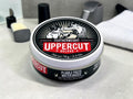 A round tin of Uppercut Featherweight Deluxe hair pomade sits on a light gray countertop with a towel and grooming items blurred in the background.