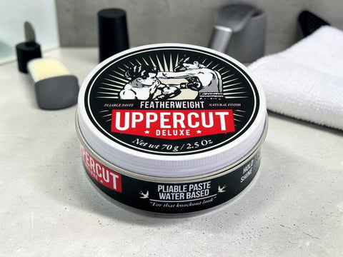 A round tin of Uppercut Featherweight Deluxe hair pomade sits on a light gray countertop with a towel and grooming items blurred in the background.