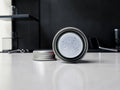 A silver metal cylindrical container with a clear plastic insert featuring multiple circular holes, shown with its lid removed and placed beside it on a light gray countertop.