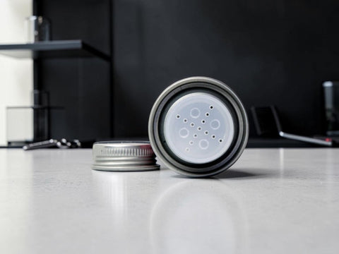 A silver metal cylindrical container with a clear plastic insert featuring multiple circular holes, shown with its lid removed and placed beside it on a light gray countertop.