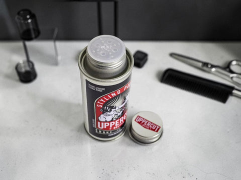 A can of Uppercut Styling Product with its cap removed, placed on a light countertop with scissors, a comb, and other grooming tools nearby.