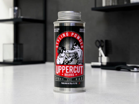 A can of Uppercut Deluxe Styling Powder standing on a light countertop in a modern kitchen or bar setting with dark shelves and bar tools blurred in the background.