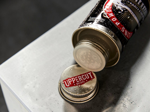 A close-up of a metal bottle cap labeled 'UPPERCUT DELUXE' resting beneath an open canister, both covered in water droplets, on a gray textured surface.